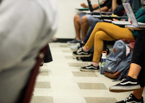 Students sitting in desks in a classroom shown from the waist down. They are in one-piece desks with casual postures.